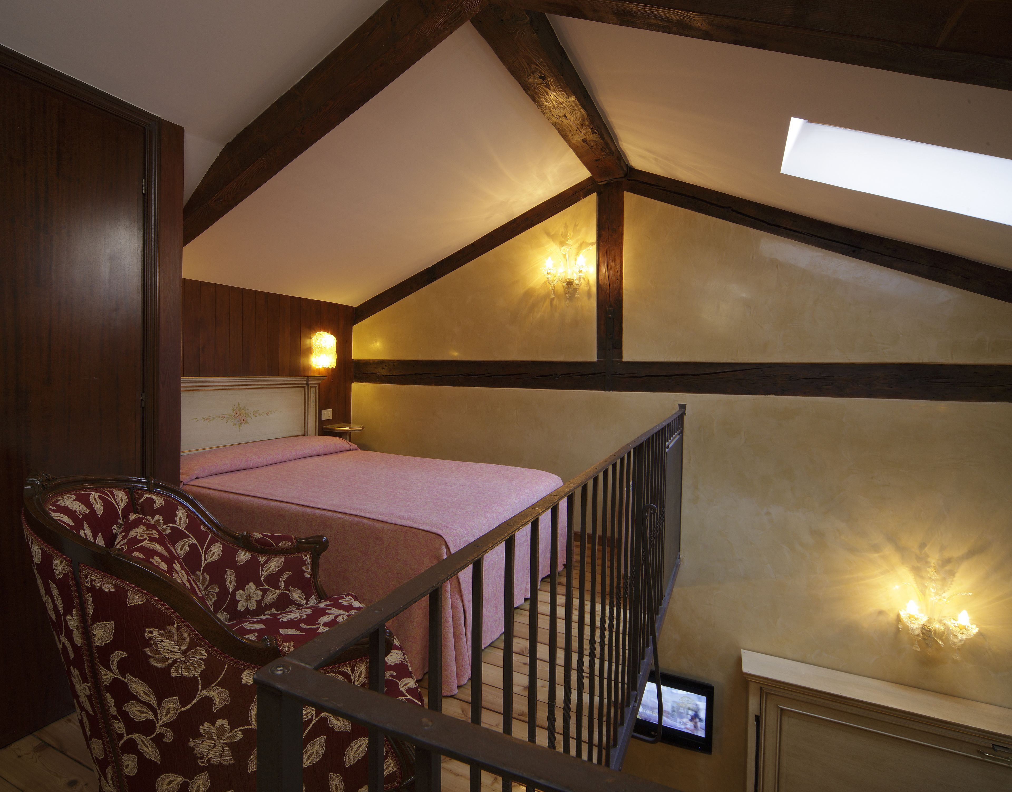 A bedroom with a sloped ceiling, a bed with a pink bedspread, a wooden chair, and a television on the wall.