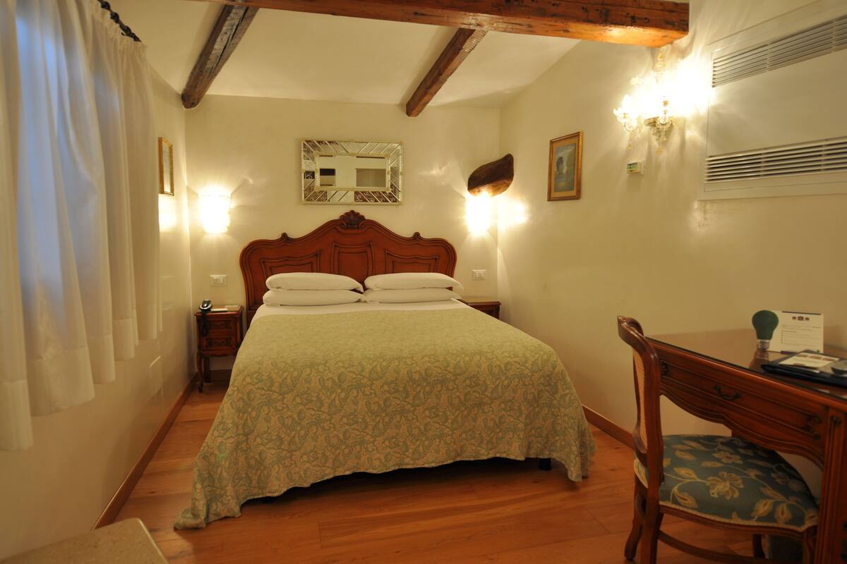 A bedroom with a wooden bed, a nightstand, a desk with a chair, and a wall-mounted mirror.