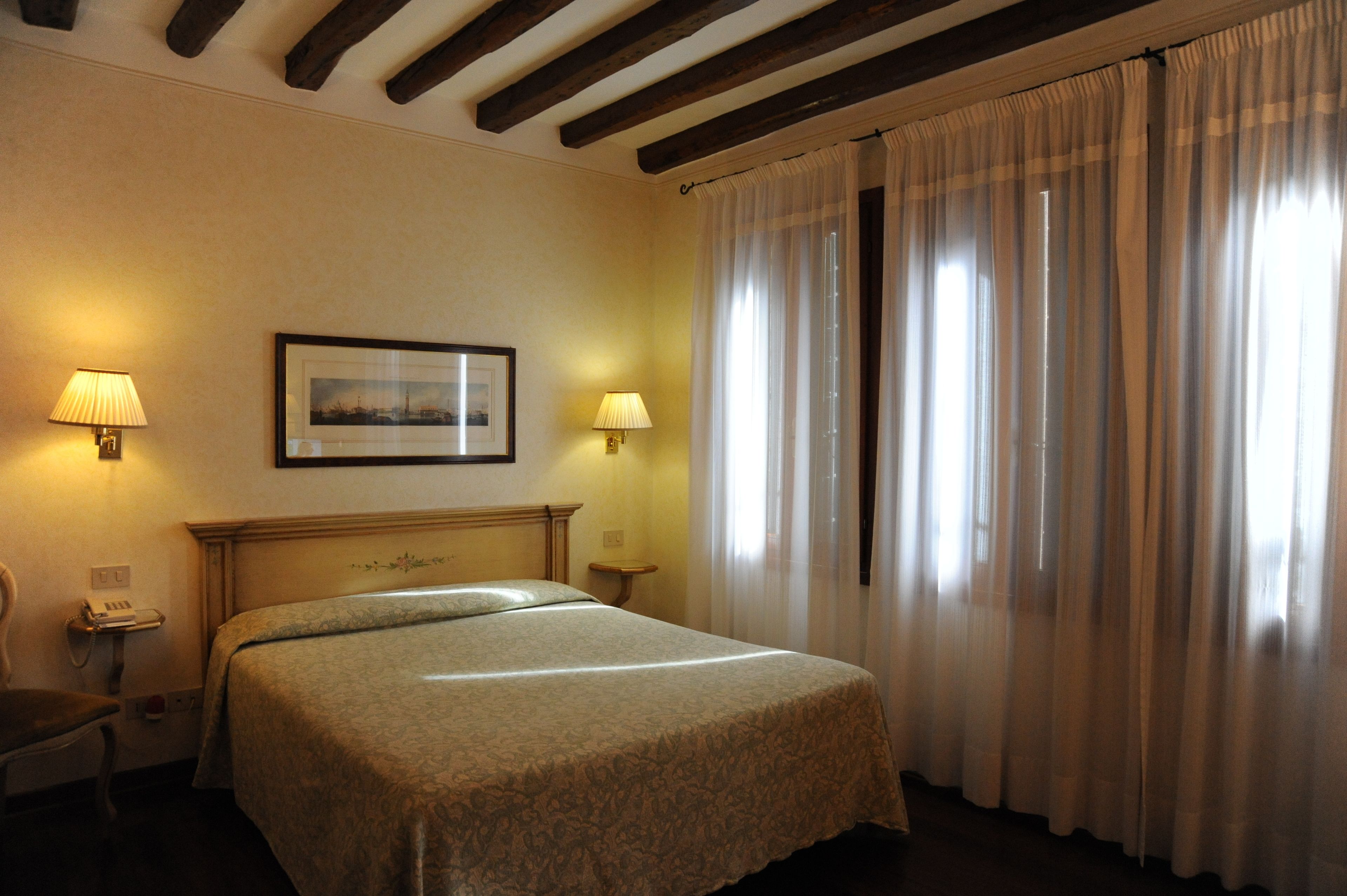 Ein Hotelzimmer mit einem Bett, zwei Nachttischlampen, einem gerahmten Bild an der Wand und einem Fenster mit Vorhängen.