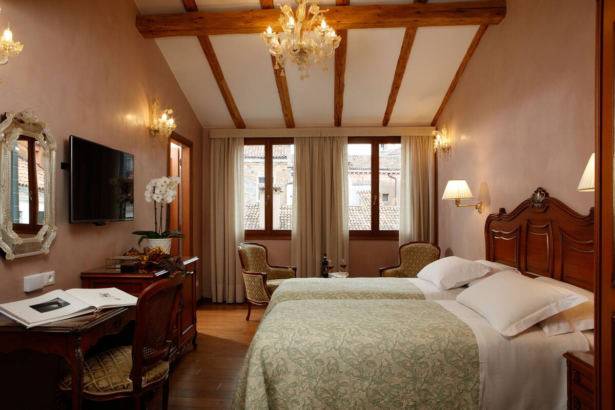 A hotel room with two beds, a desk with a chair, a TV, and a chandelier.