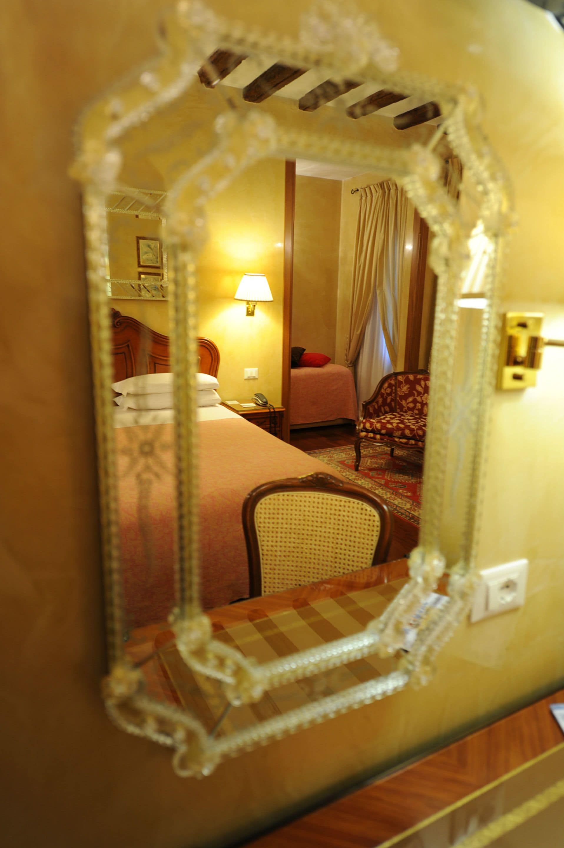 A mirror reflecting a hotel room with a bed, a chair, a nightstand, and a lamp.