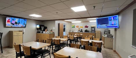 A dining area with multiple tables and chairs, a kitchen counter, and two televisions mounted on the walls.