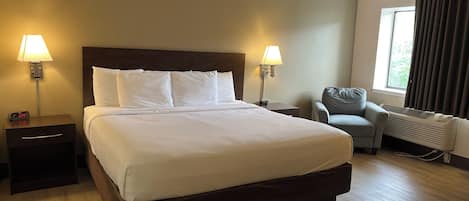 Una habitación de hotel con una cama grande, dos mesitas de noche con lámparas, una silla y un ventanal con cortinas.