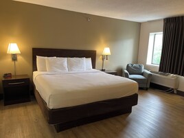 Una habitación de hotel con una cama grande, dos mesitas de noche con lámparas, una silla y un ventanal con cortinas.