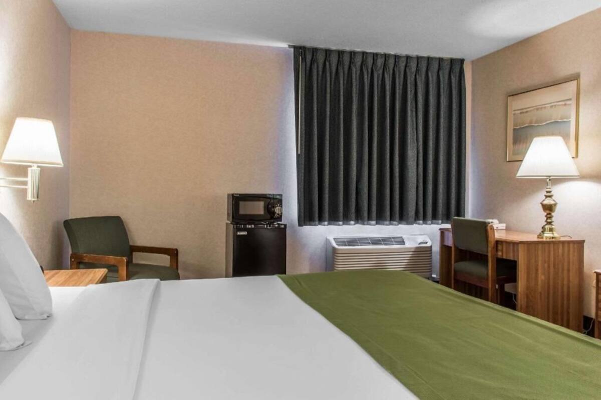 A hotel room with a bed, two chairs, a desk, a microwave, and an air conditioning unit.