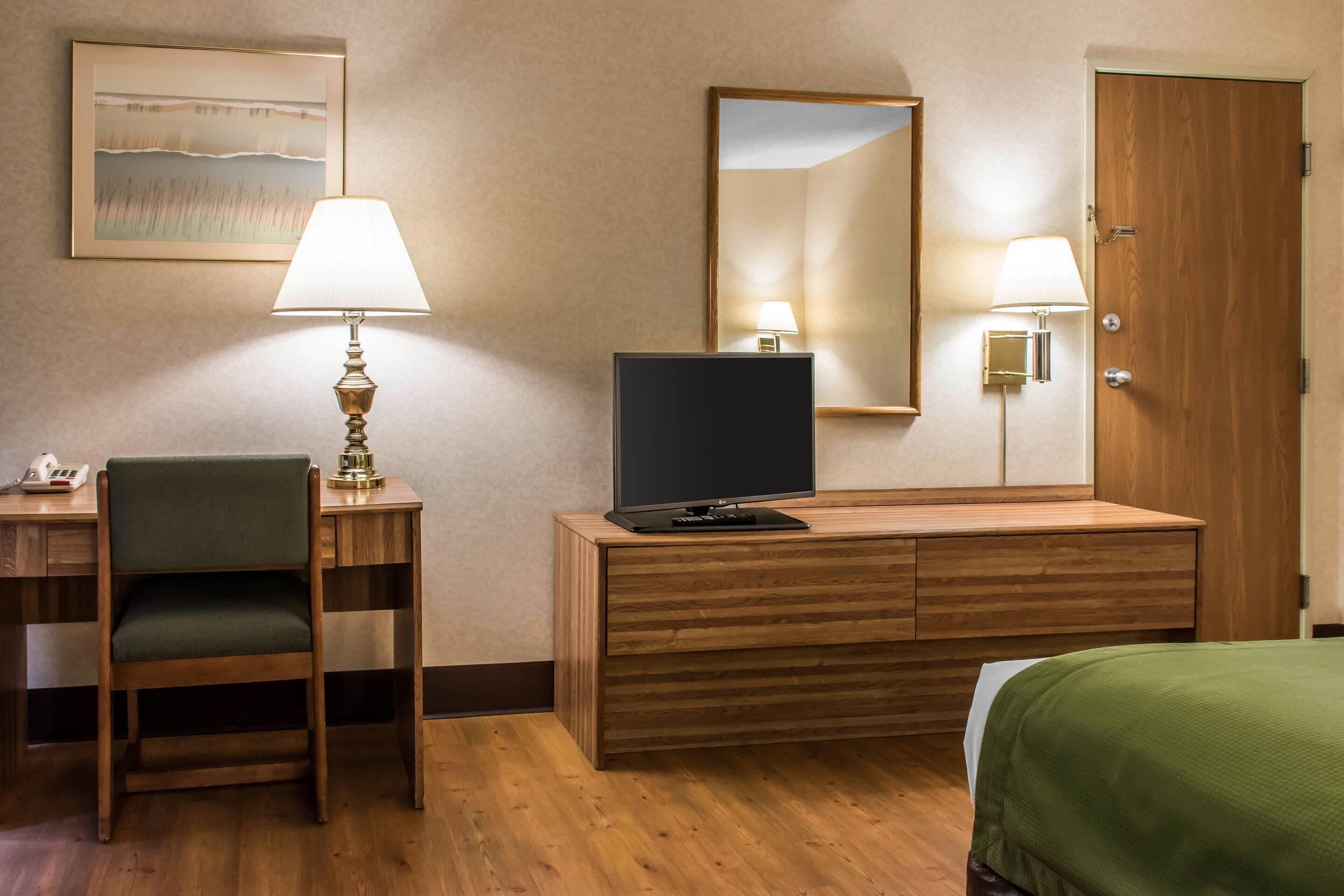 A hotel room with a wooden desk, a chair, a television, a bed with a green bedspread, a wall-mounted mirror, and two wall lamps.