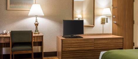 A hotel room with a wooden desk, a chair, a television, a bed with a green bedspread, a wall-mounted mirror, and two wall lamps.