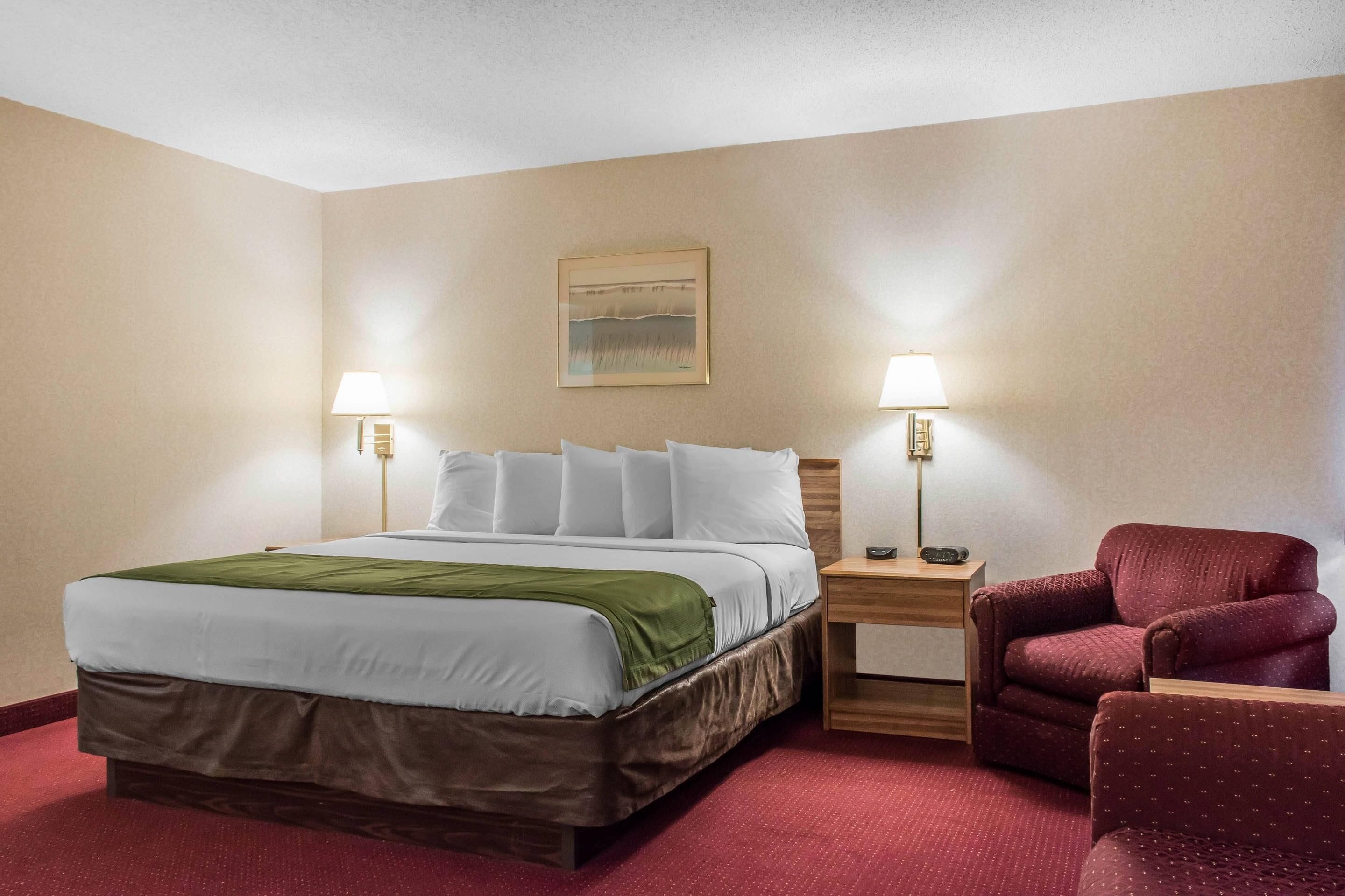 A hotel room with a large bed, two bedside tables with lamps, a red armchair, and a painting on the wall.