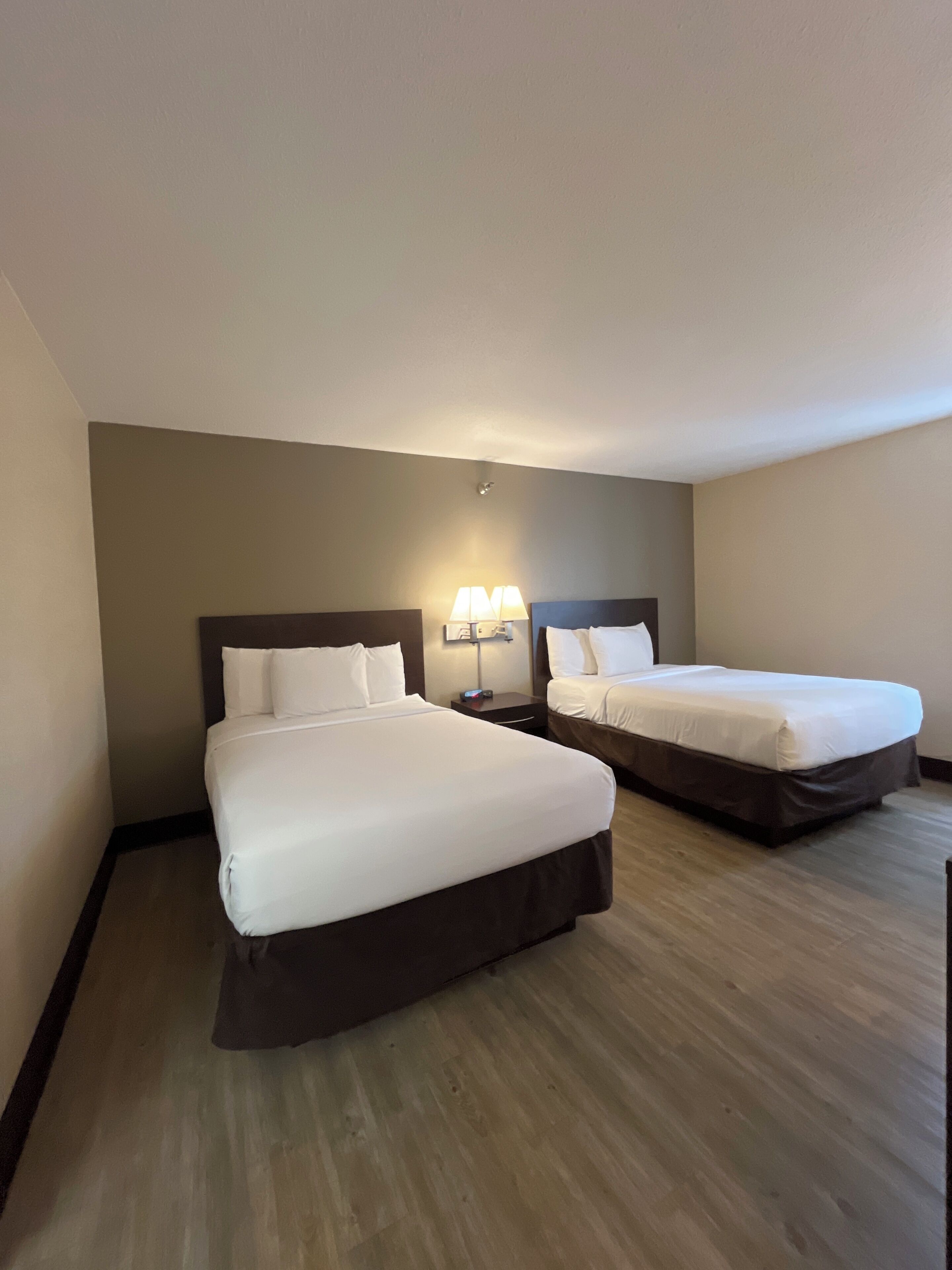 A hotel room with two beds, a nightstand with a lamp, and a wooden floor.