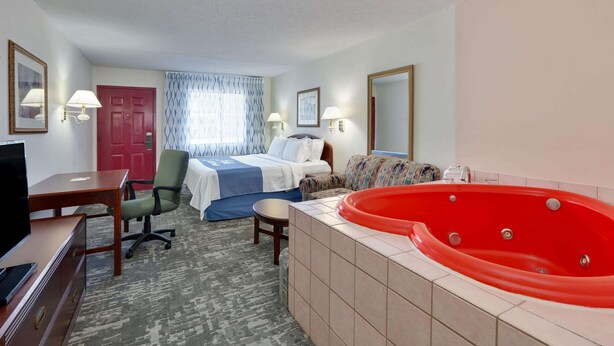 A hotel room with a bed, a red jacuzzi, a desk with a TV, a chair, a small table, and a carpeted floor.
