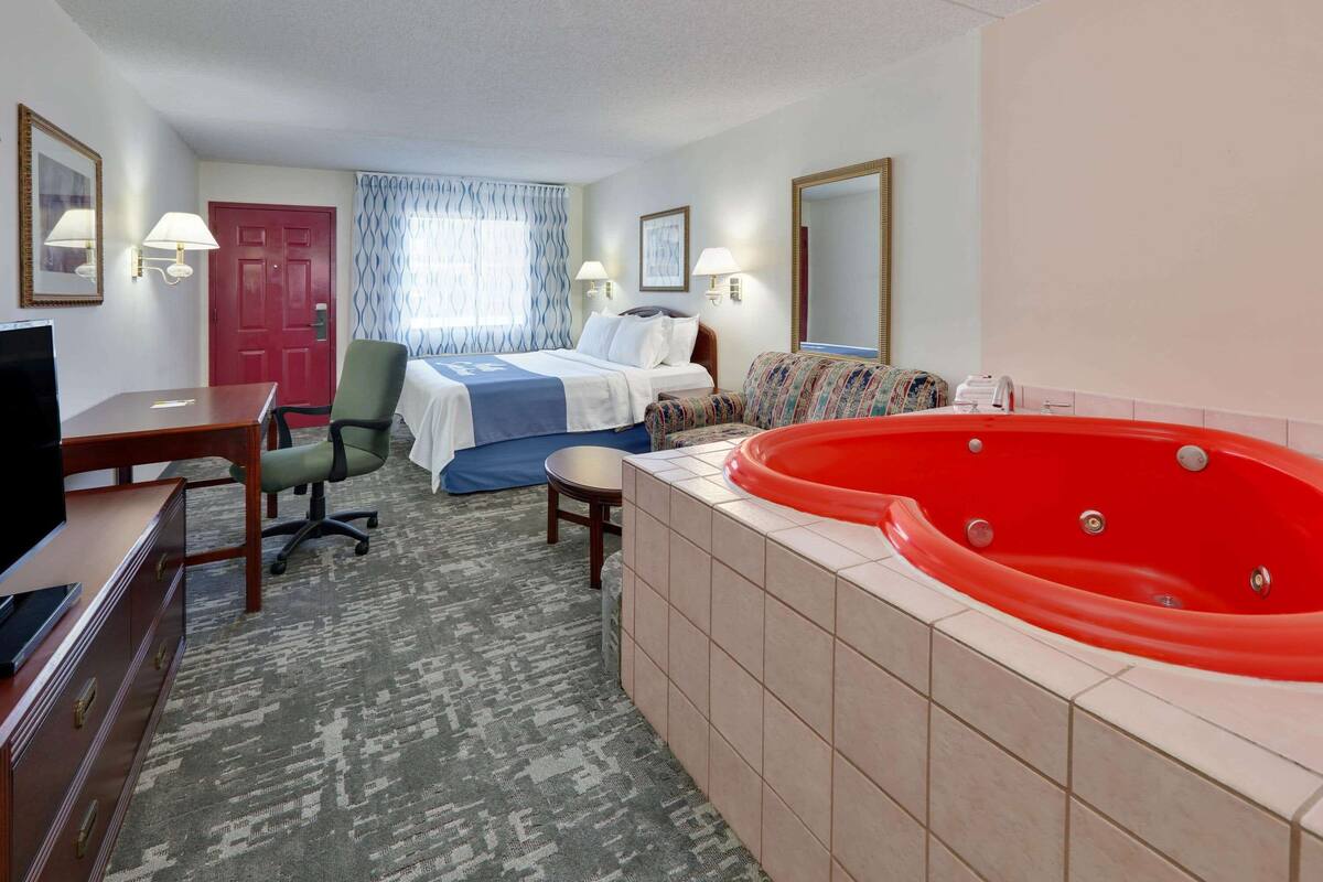 Een hotelkamer met een bed, een rode jacuzzi, een bureau met een televisie, een stoel, een klein tafeltje en een vloerbedekking.