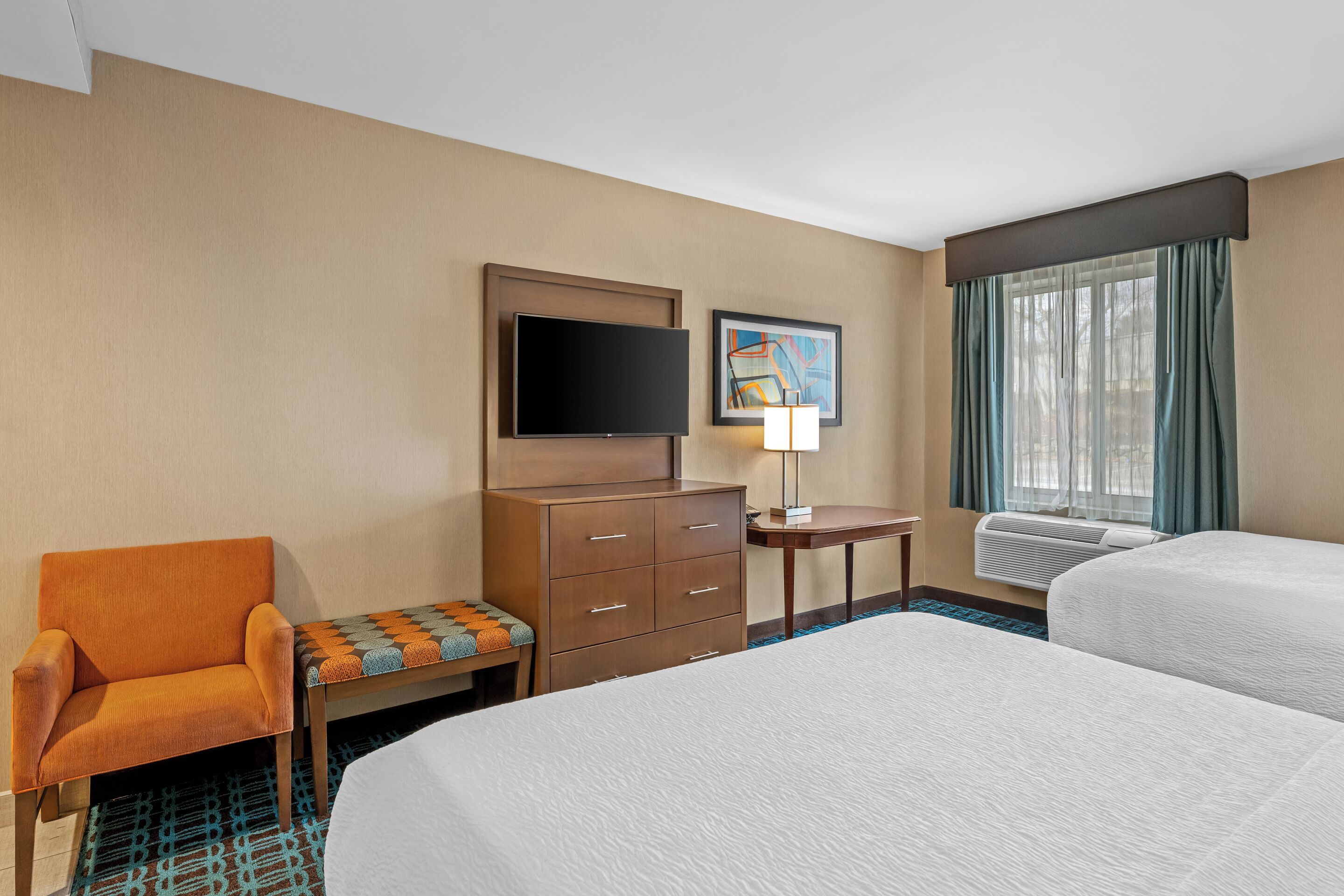 A hotel room with two beds, a TV, a desk with a lamp, a chair, and a window with curtains.