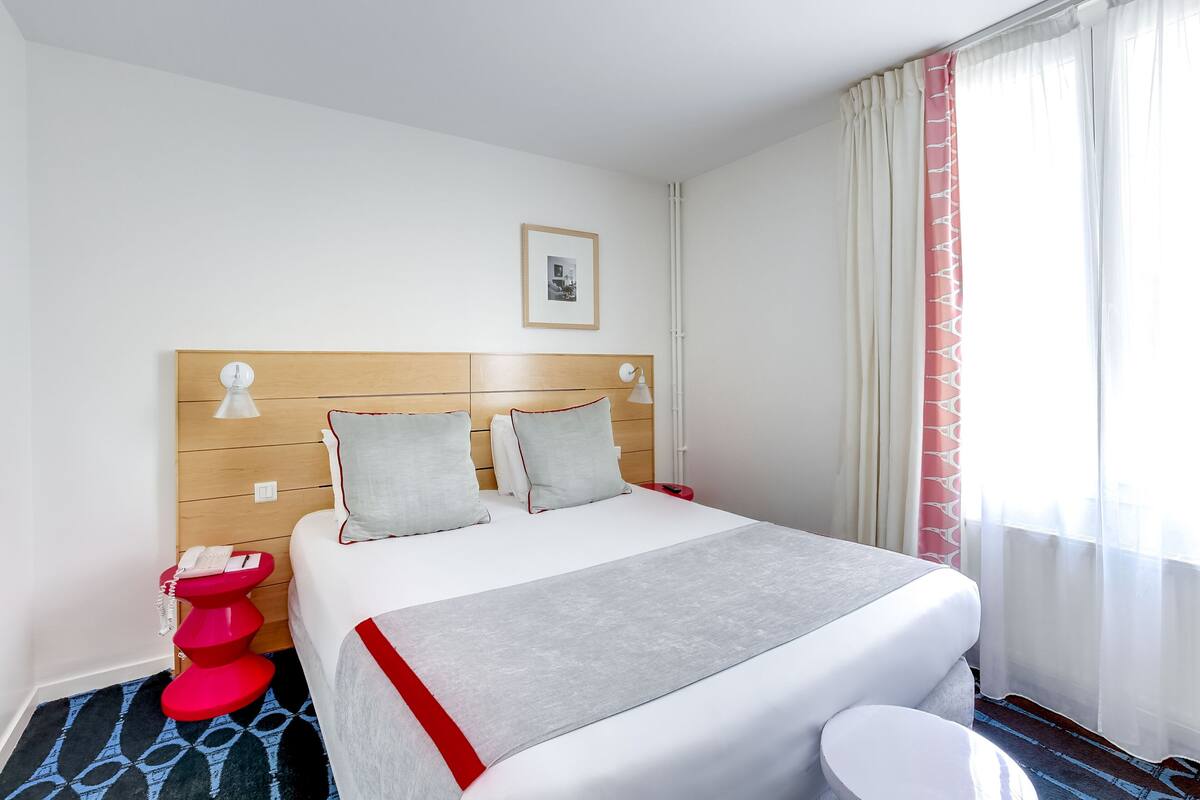 Habitación de hotel con cabecera de madera, cama con ropa blanca, mesita de noche roja con teléfono y una ventana con cortinas traslúcidas.