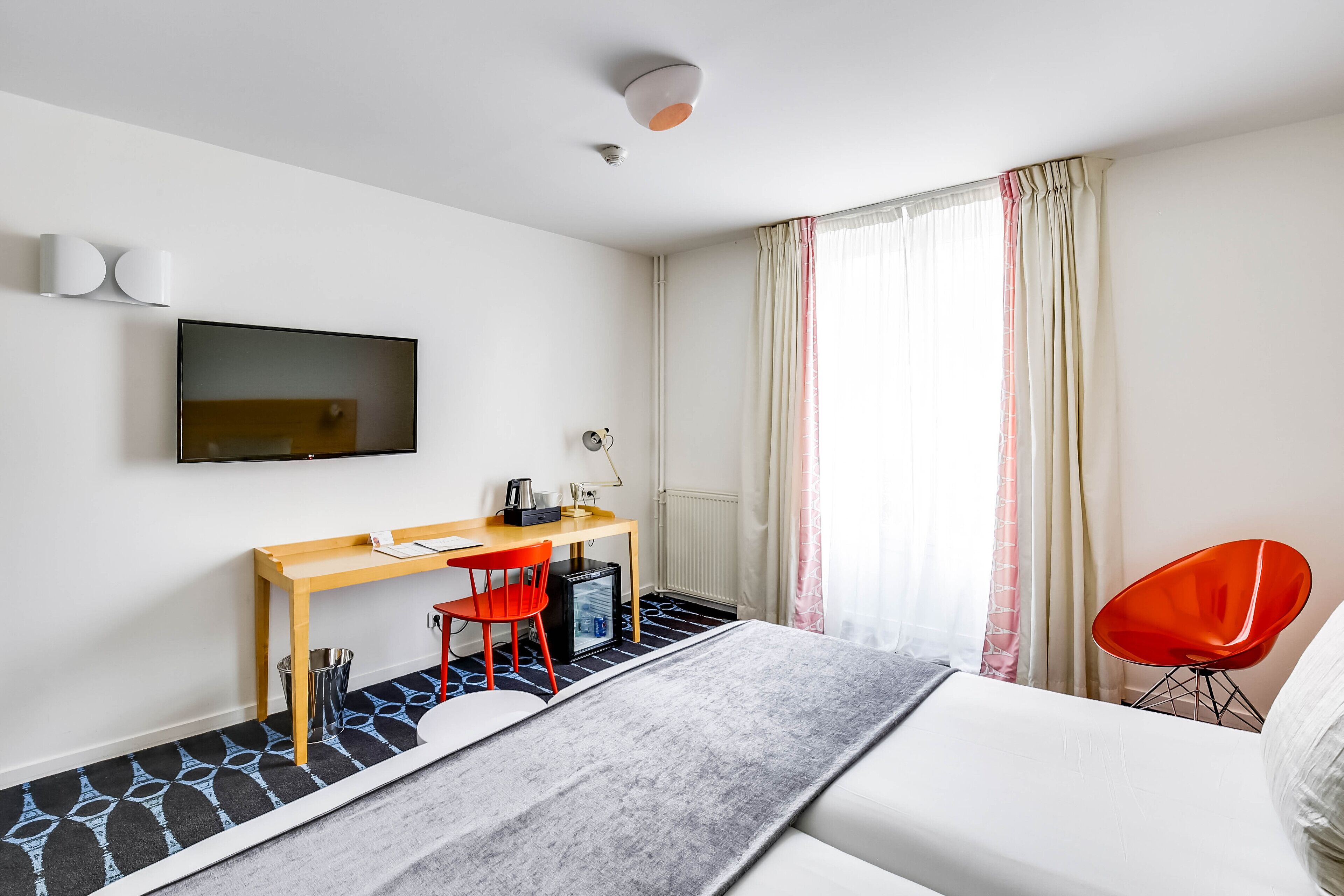 A modern hotel room with a bed, a desk with a computer, a chair, and a TV on the wall.