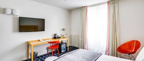 A modern hotel room with a bed, a desk with a computer, a chair, and a TV on the wall.