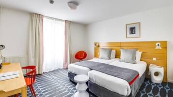 A hotel room with a bed, a desk, a chair, a bedside table, and a window with curtains.