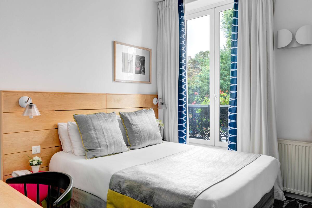 Habitación de hotel ordenada con cabecera de madera, cama con ropa de cama gris y amarilla, una mesita con una silla y una ventana con cortinas estampadas.