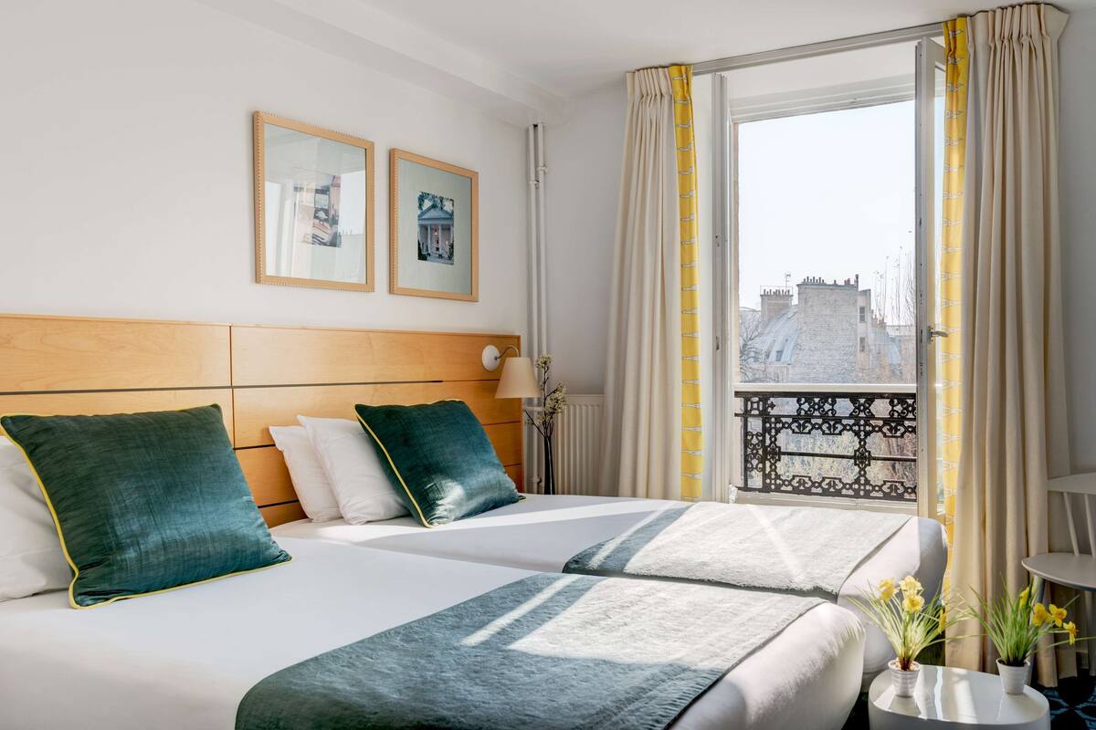 Habitación de hotel con dos camas, cabecera de madera, ventana con cortinas, una mesita con un florero y dos cuadros en la pared.
