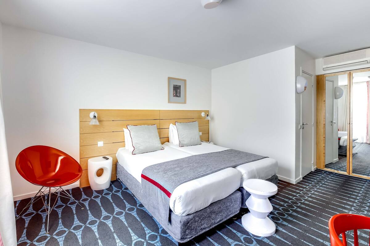 Una habitación de hotel con cama, una silla roja, un inodoro blanco y un espejo.