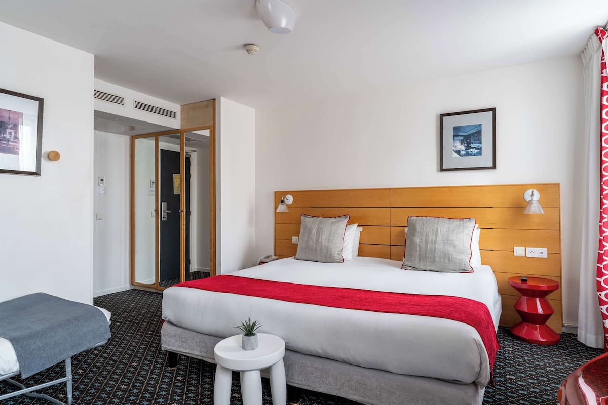Habitación de hotel con una cama grande, una mesita con una planta, una colcha roja y blanca, y un cuadro en la pared.