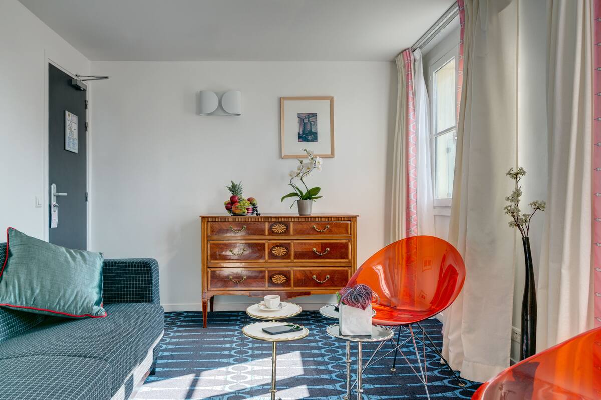 Una habitación con alfombra azul estampada, un aparador de madera con cajones, una silla roja, una mesita con taza y platillo, y un cuadro enmarcado en la pared.