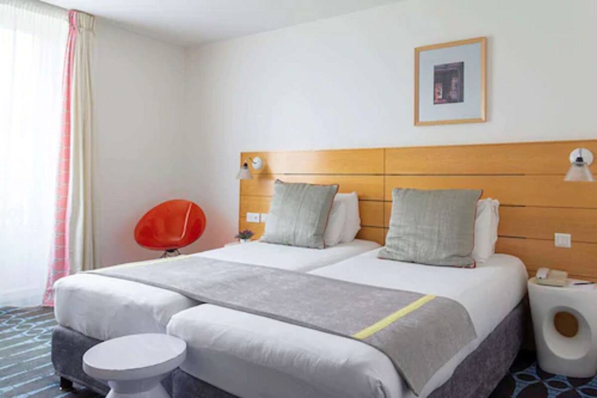 Habitación de hotel con dos camas, una silla roja, una mesita redonda y un cuadro enmarcado en la pared.