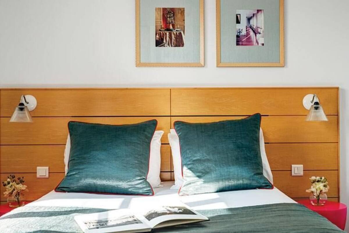 Una cama con cabecera de madera, dos almohadas, una mesita de noche y dos lámparas fijadas a la pared.