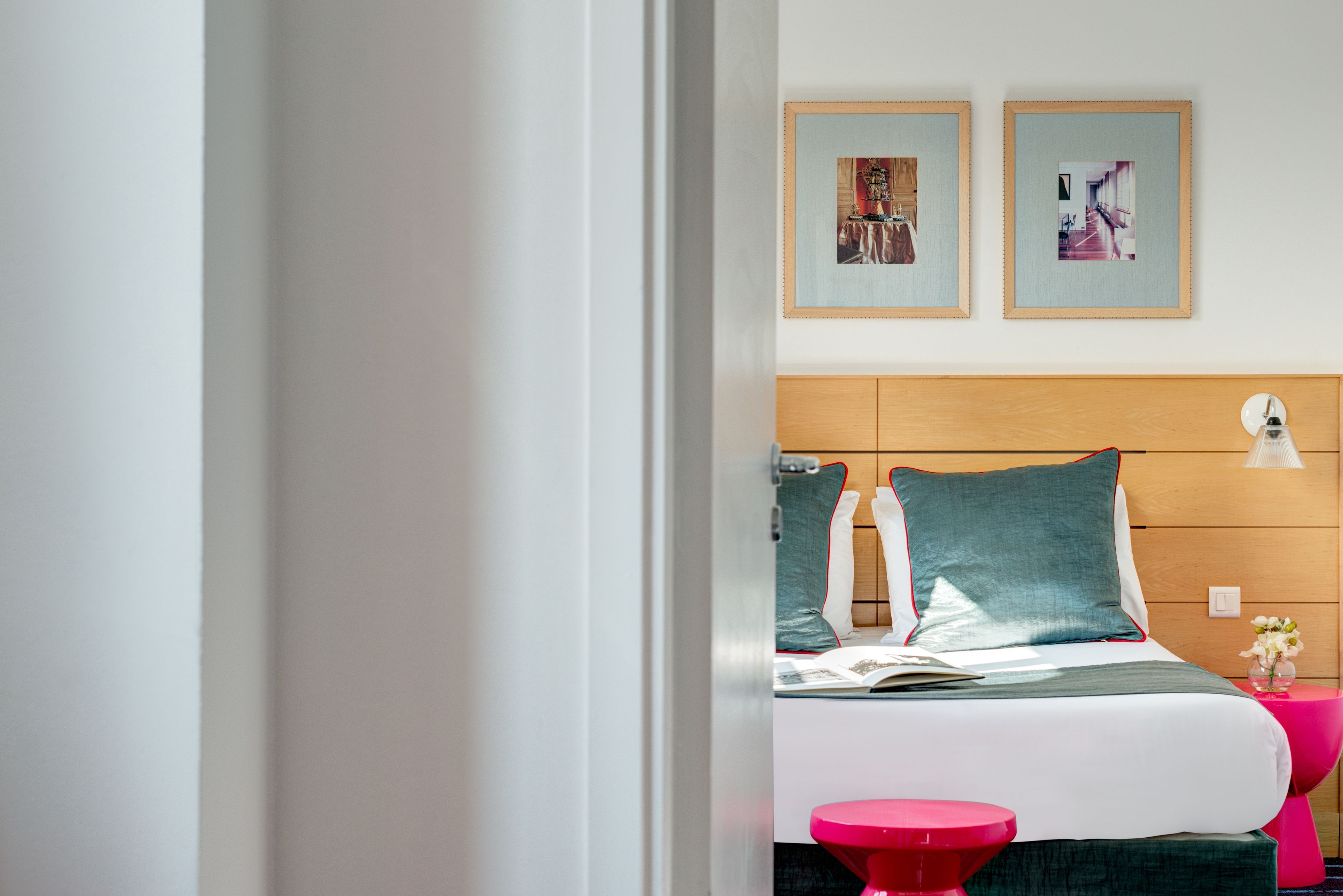 Ein Hotelzimmer mit einem Holzkopfteil, einem Bett mit türkisfarbener und weißer Bettwäsche, zwei gerahmten Bildern an der Wand und einem kleinen Tisch mit einer pinken Lampe.