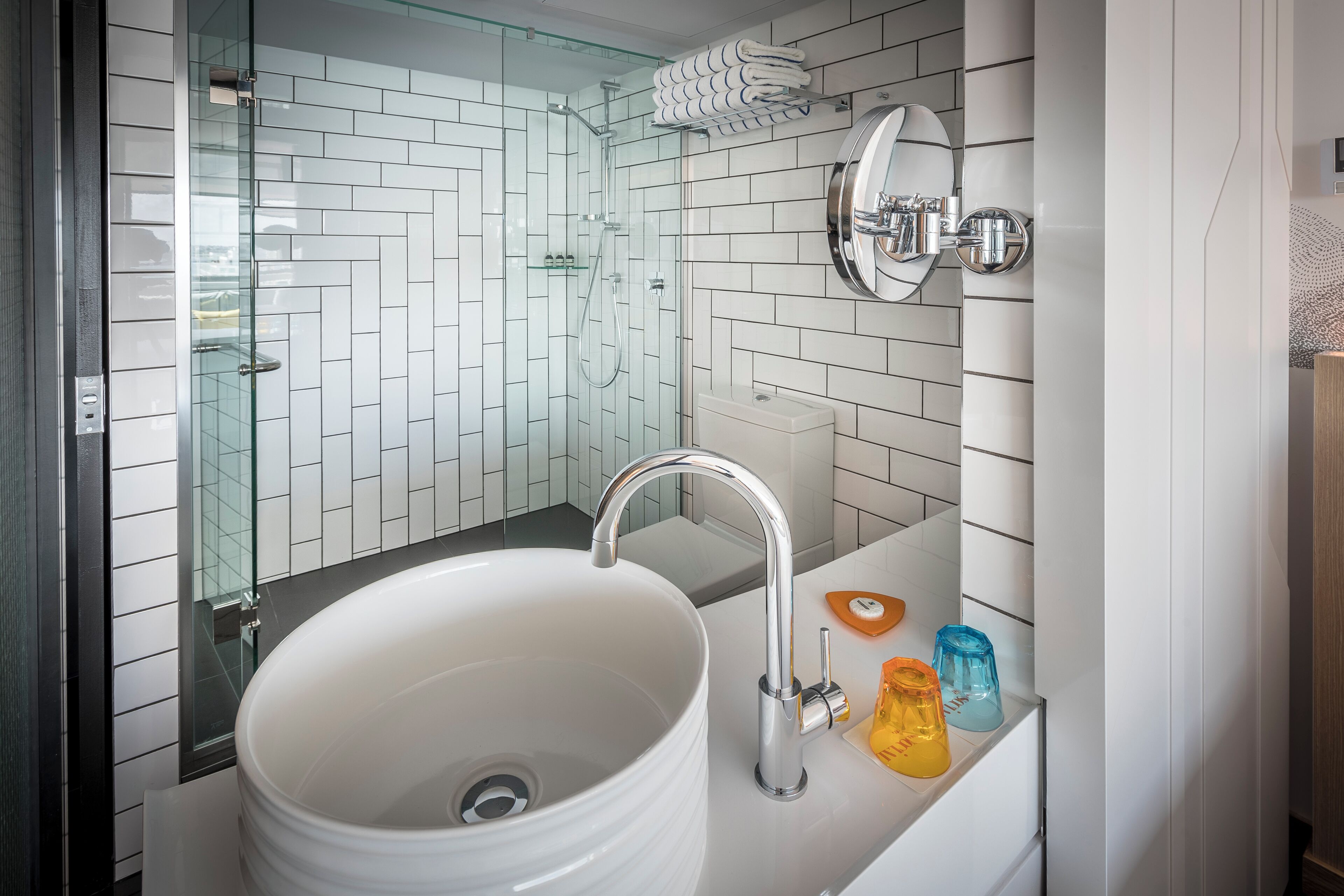 Un baño moderno con un lavamanos blanco, cabina de ducha con paredes de vidrio y un espejo con marco redondo.