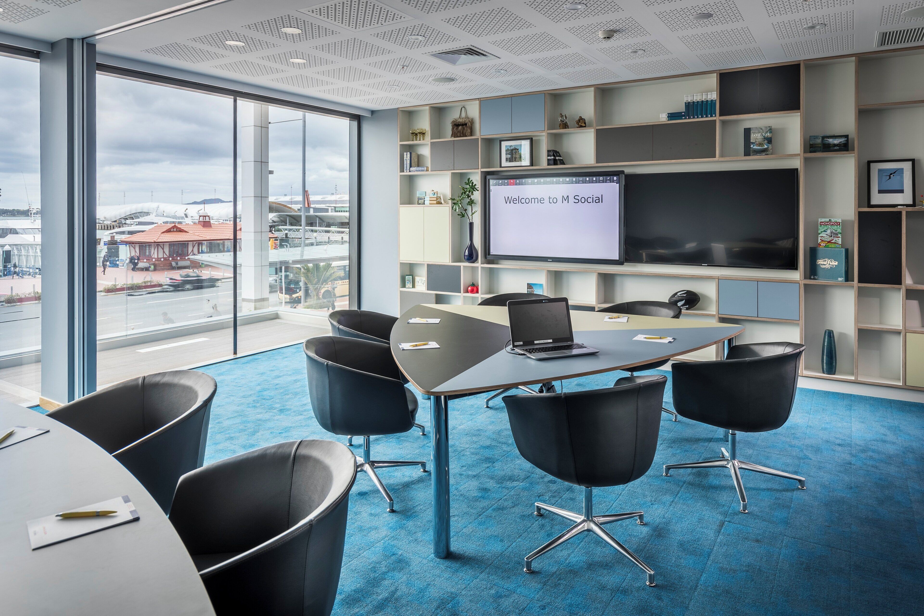 Une salle de conférence moderne dotée d’un grand écran affichant le message « Bienvenue à M Social », d’un sol recouvert d’une moquette bleue et de chaises noires disposées autour d’une table.