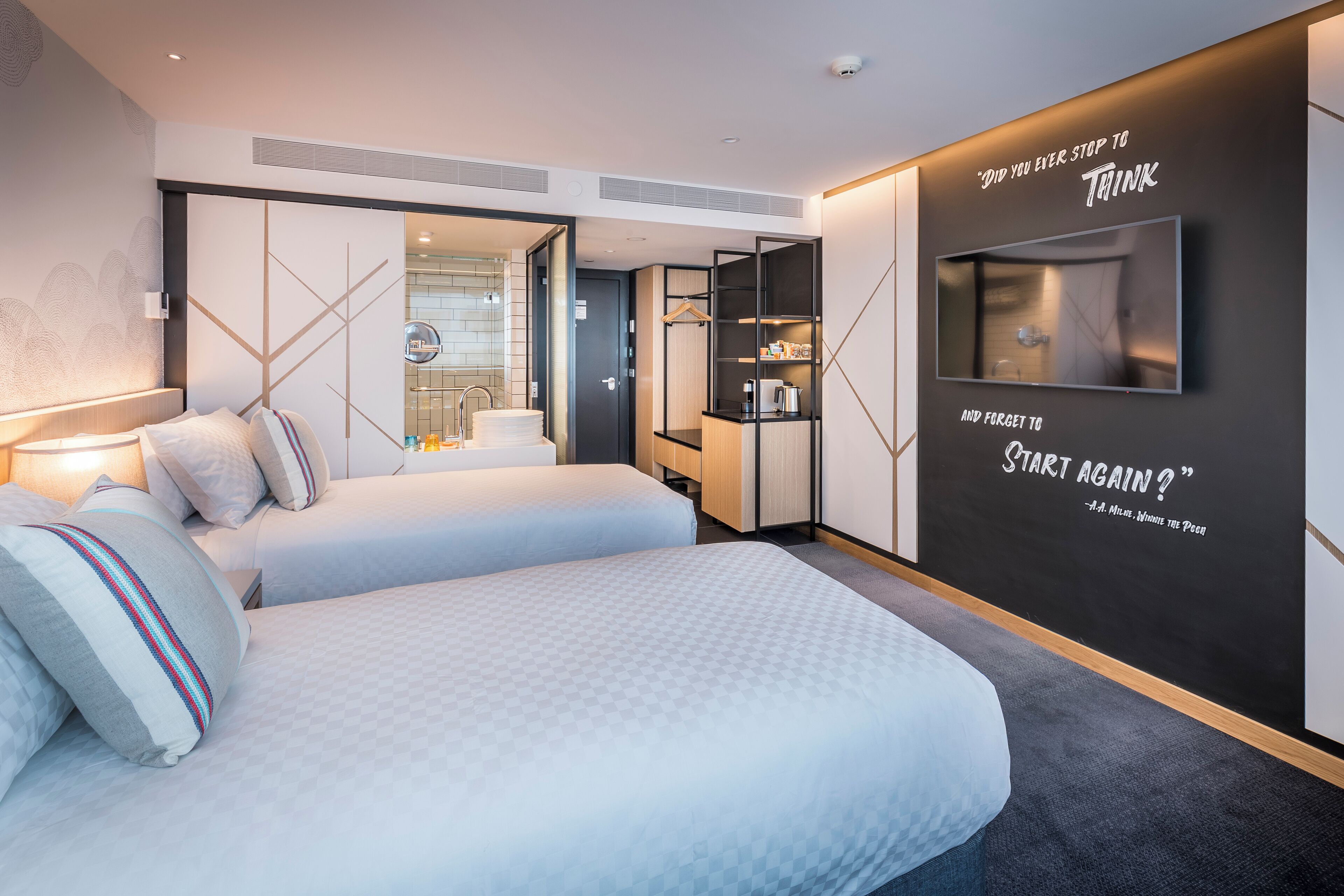 A modern hotel room with a large bed, a TV, a mirror, and a motivational quote on the wall.