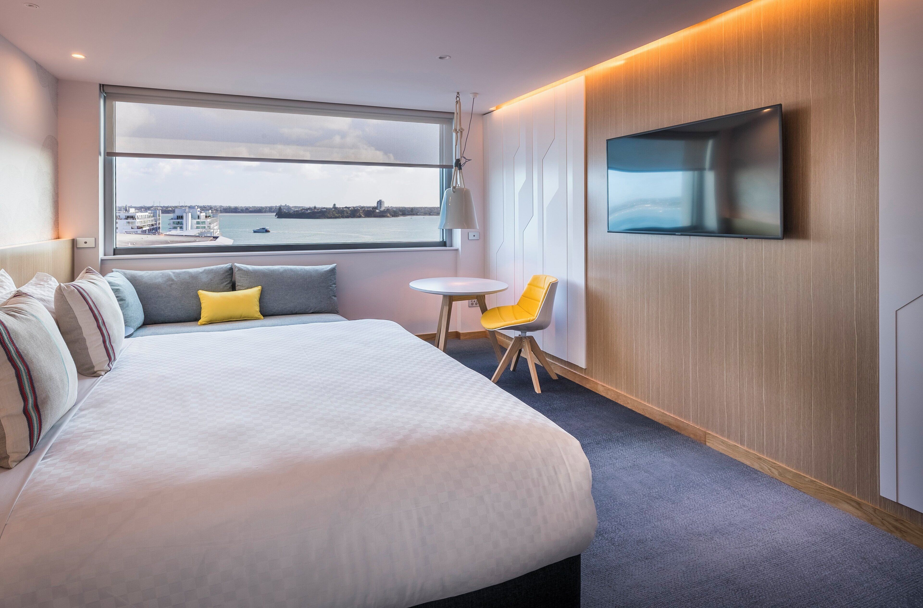 Een moderne hotelkamer met een groot bed, een klein rond tafeltje, een gele stoel, een aan de muur bevestigde televisie en uitzicht op de stad door een groot raam.
