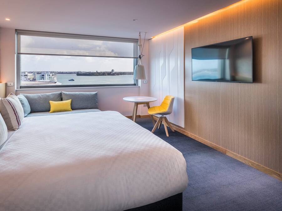Een moderne hotelkamer met een groot bed, een klein rond tafeltje, een gele stoel, een aan de muur bevestigde televisie en uitzicht op de stad door een groot raam.