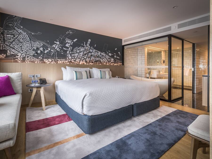 Habitació d'hotel moderna amb un llit gran, un mural de paisatge urbà, un bany amb dutxa de vidre i una taula petita amb un telèfon i un carregador.