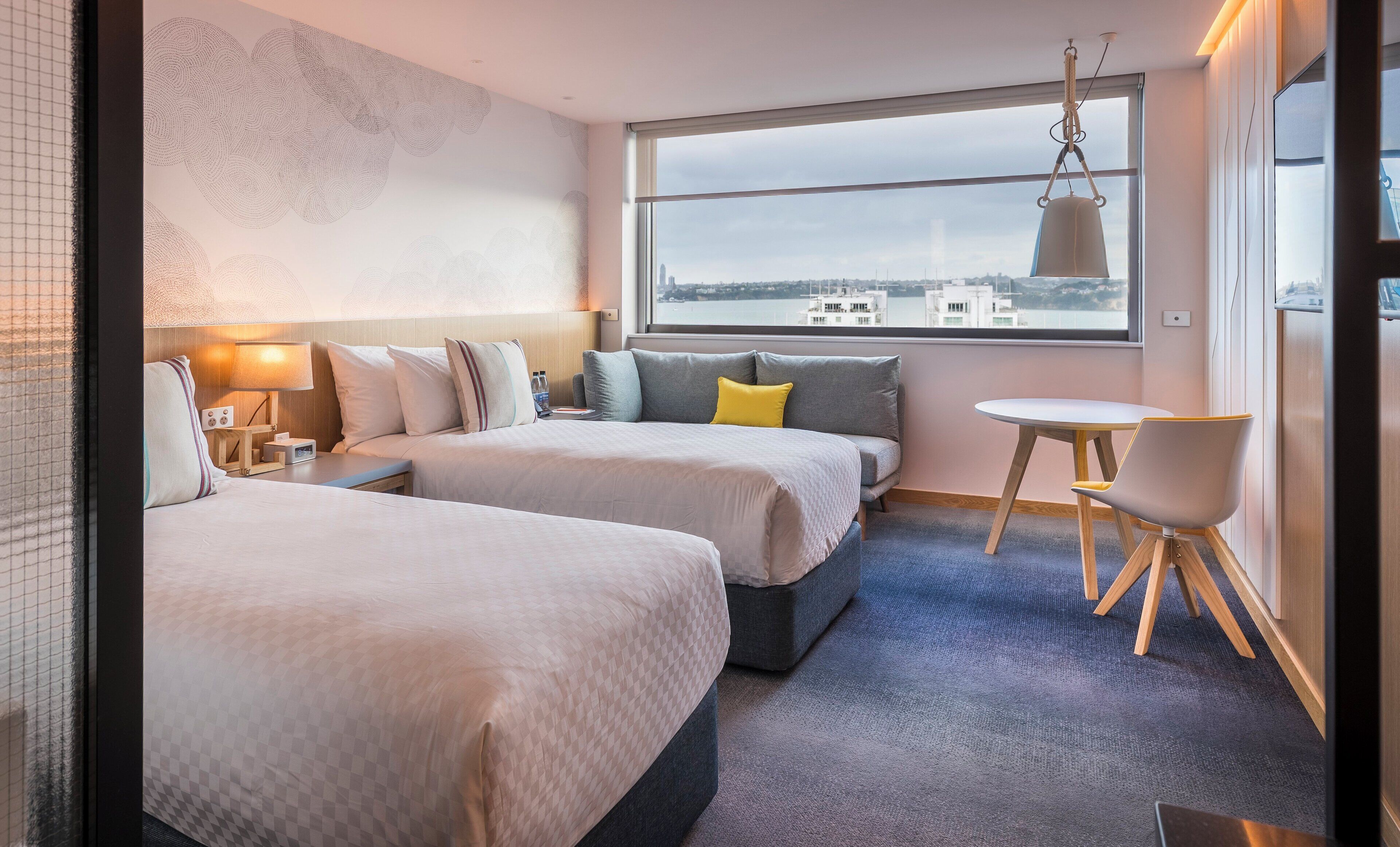 A hotel room with two beds, a small table, and a chair. There is a window with a view of buildings and a cityscape.