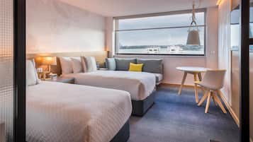 A hotel room with two beds, a small table, and a chair. There is a window with a view of buildings and a cityscape.