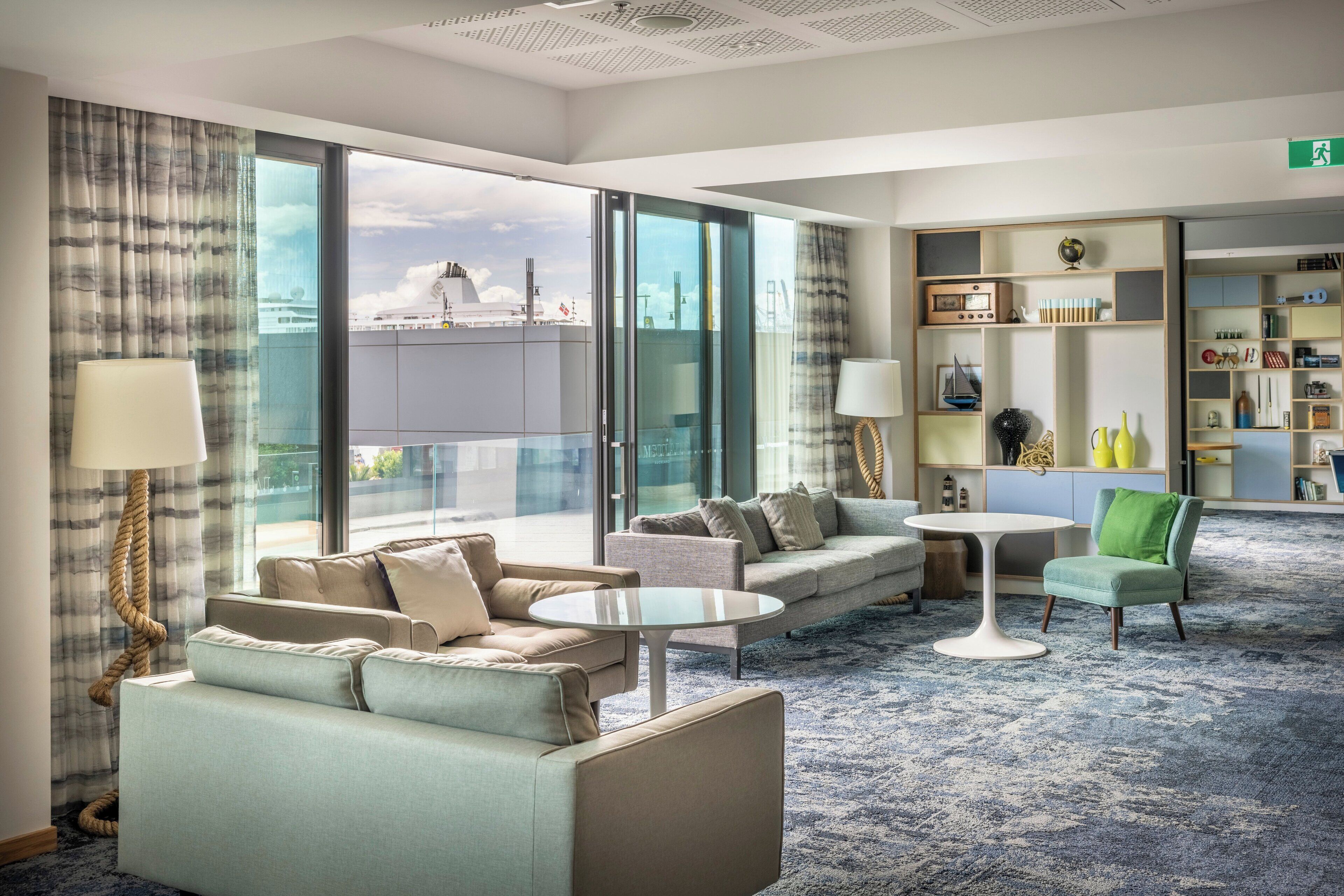 Ein modernes Hotelzimmer mit großem Fenster, einer Couch, einem Couchtisch, einem Lesesessel und einem Bücherregal.