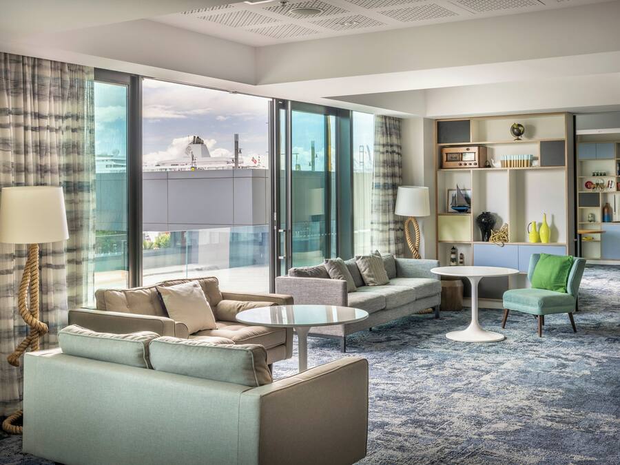 Ein modernes Hotelzimmer mit großem Fenster, einer Couch, einem Couchtisch, einem Lesesessel und einem Bücherregal.