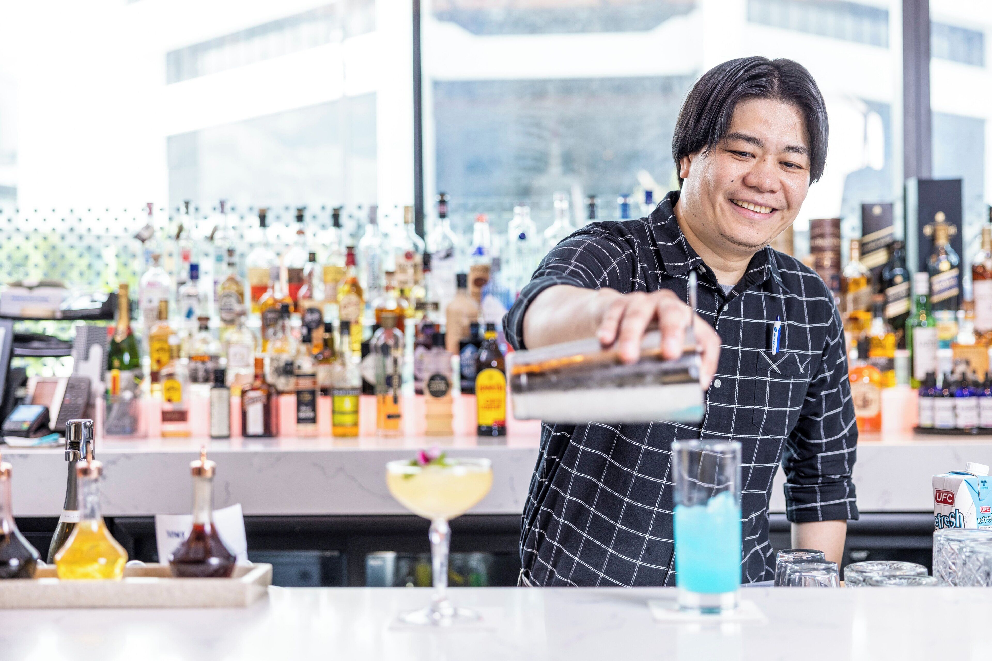 A bartender preparing a drink at a bar with various bottles and a large window in the background.