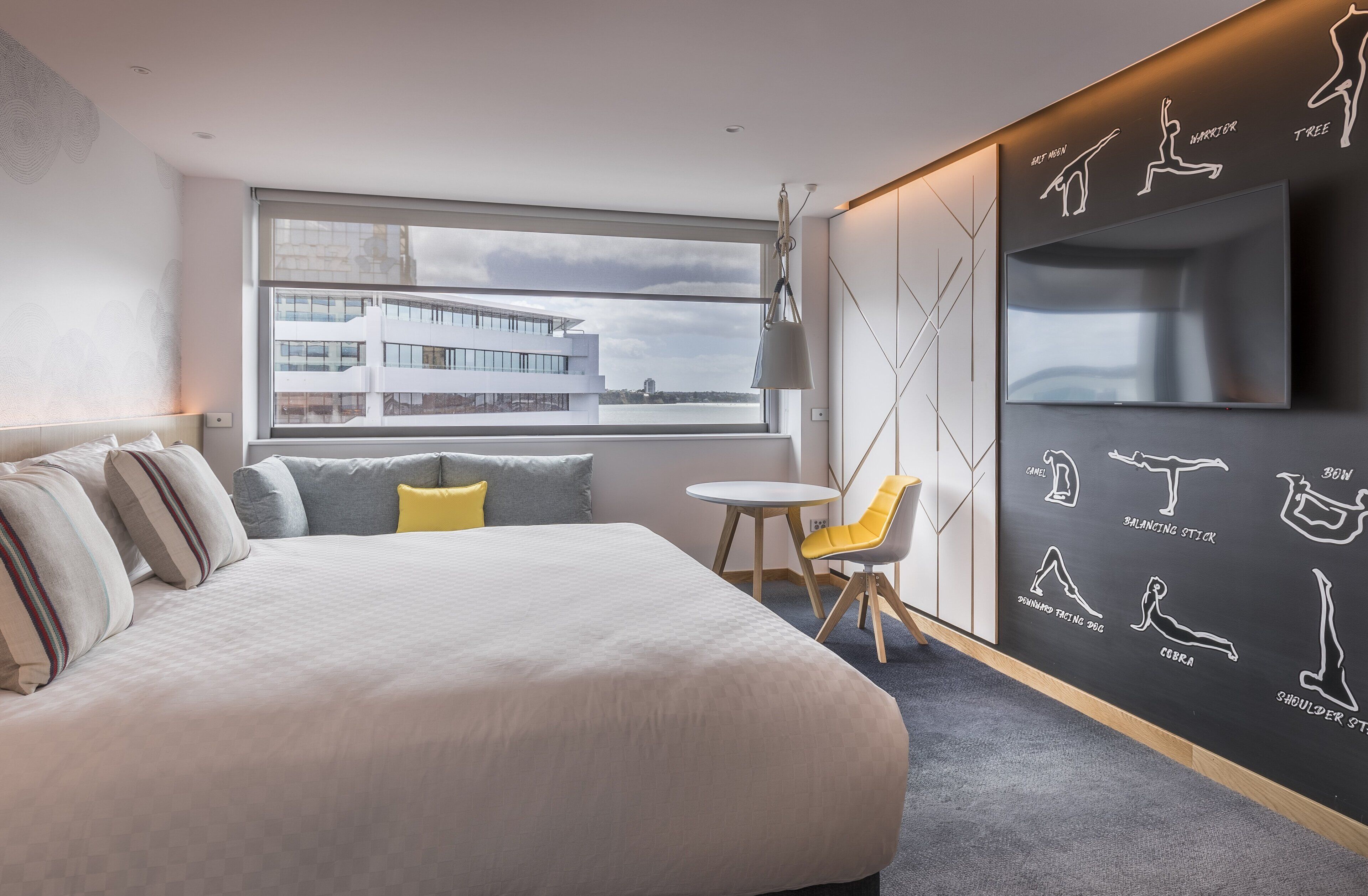 A modern hotel room with a large bed, a small table, a chair, a TV, and a wall with exercise illustrations.