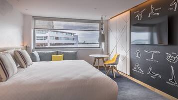 A modern hotel room with a large bed, a small table, a chair, a TV, and a wall with exercise illustrations.