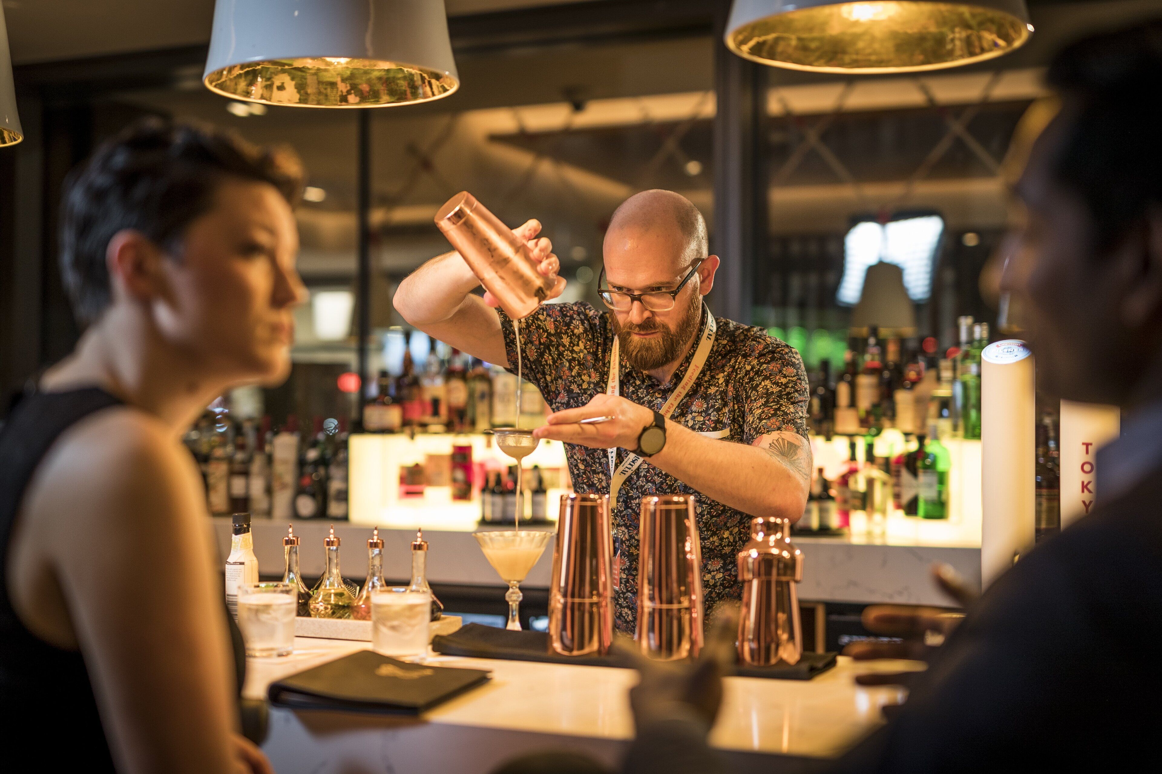 Un barman prépare un cocktail dans un bar, avec diverses bouteilles et un menu visible en arrière-plan.