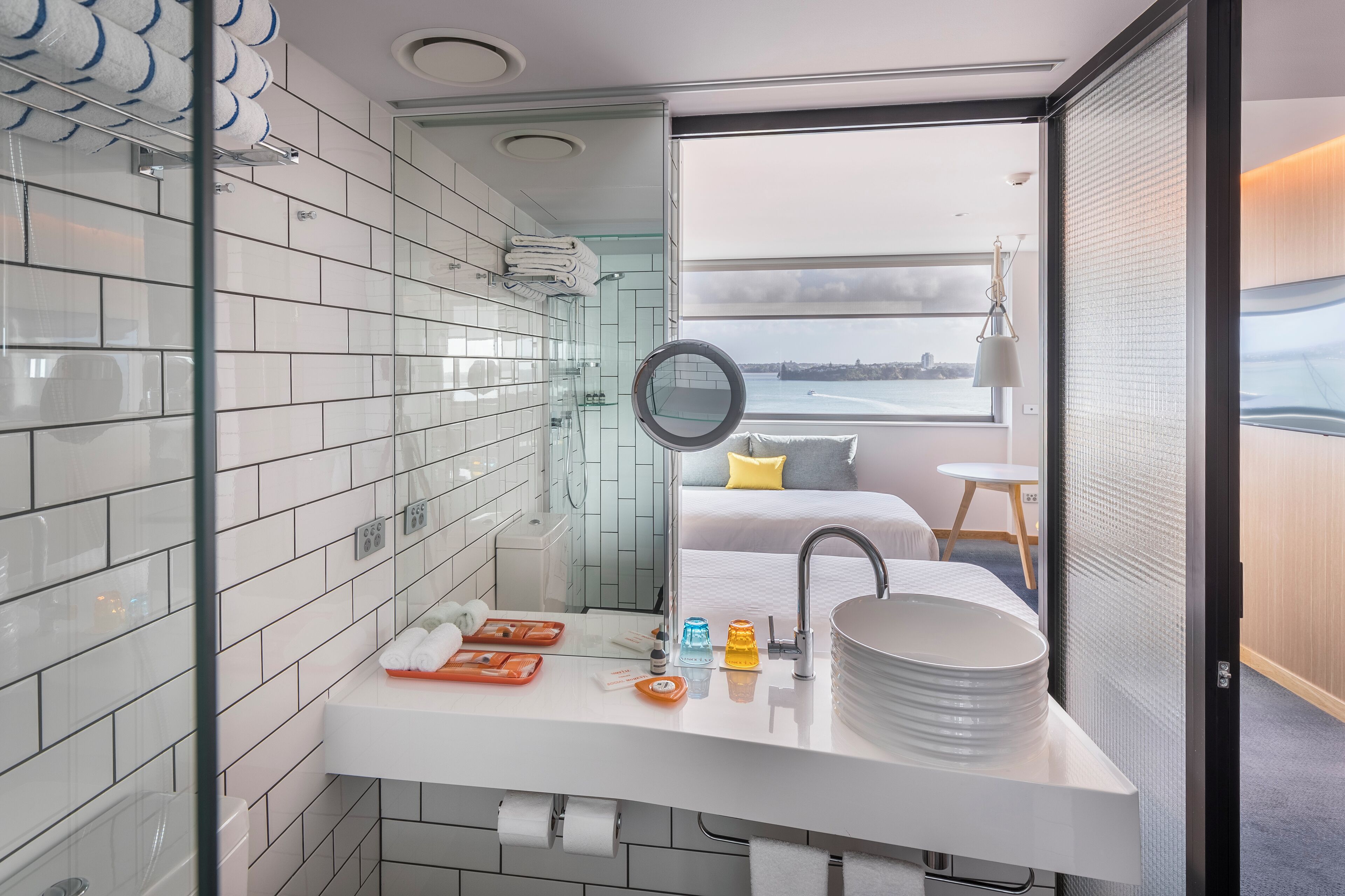 A modern bathroom with a white sink, glass shower enclosure, and a view of a bed with a yellow pillow.