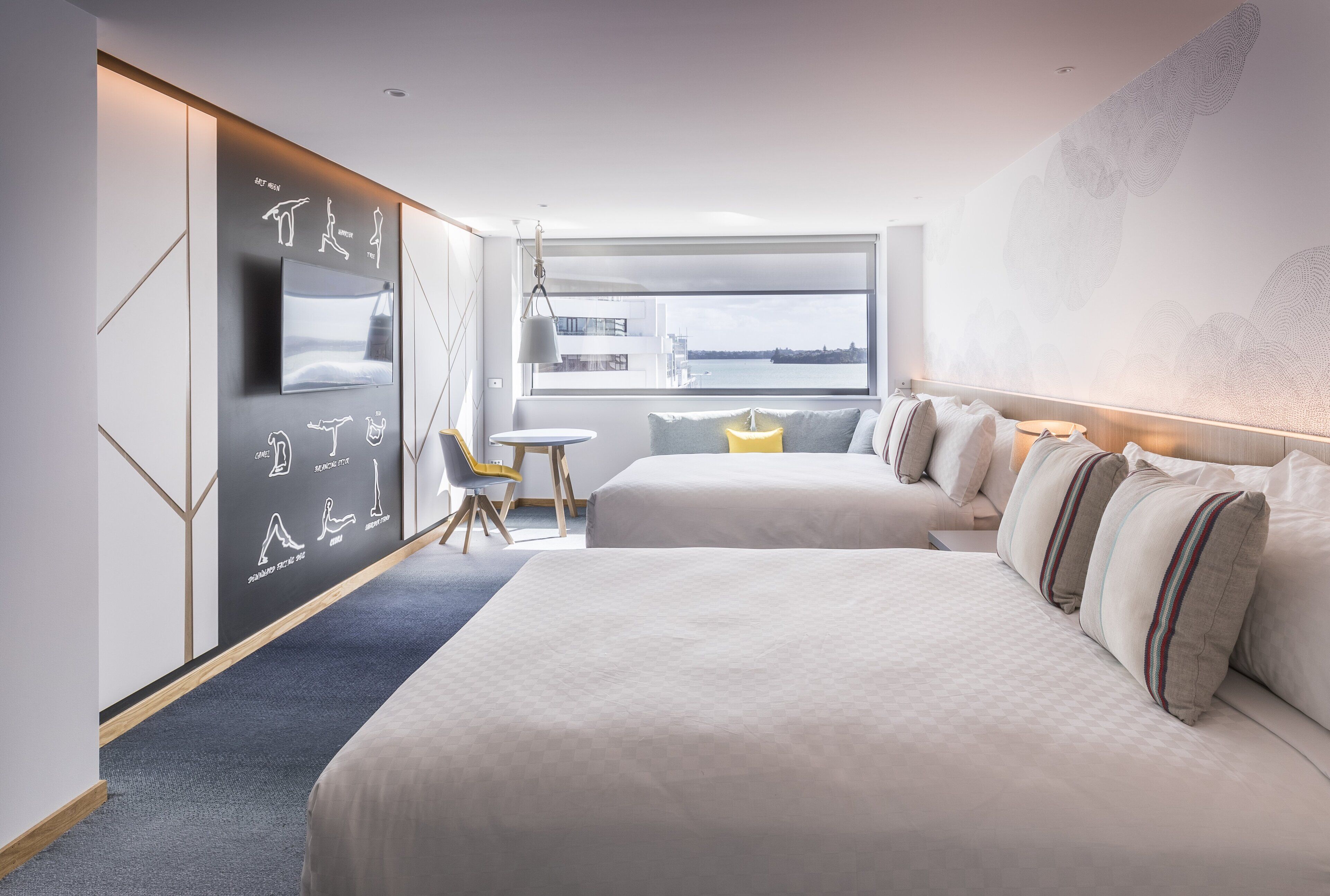 A modern hotel room with two beds, a desk, and a large window offering a view of the city.