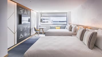 A modern hotel room with two beds, a desk, and a large window offering a view of the city.