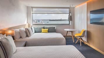 A hotel room with two beds, a small table, a chair, a TV, and a large window with a view of the water.