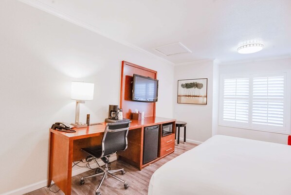 A hotel room with a wooden desk, a television, a bed, and a window with blinds.