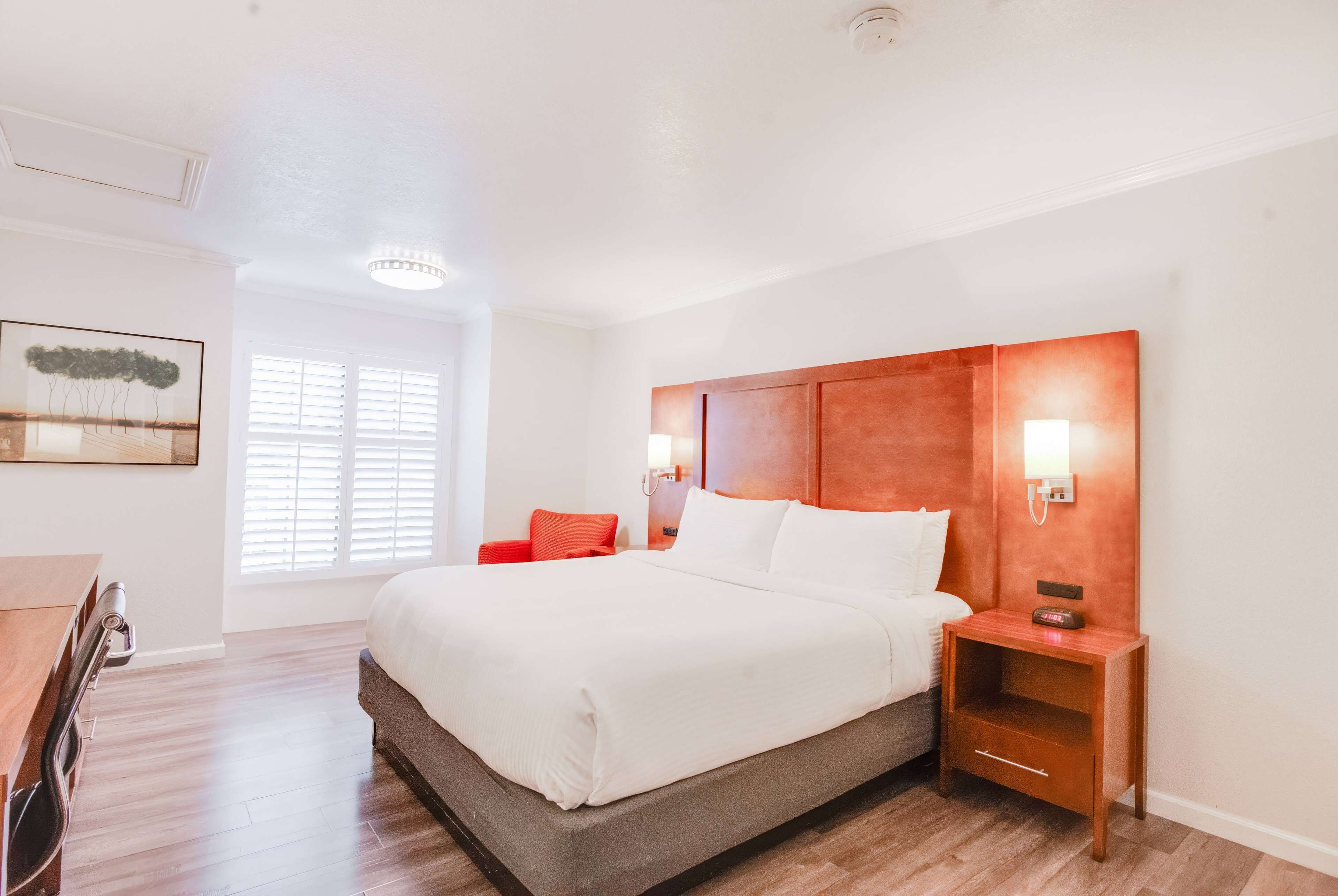 Una habitación de hotel con una cama grande, una mesita de noche de madera, un escritorio y una silla. Hay una ventana con persianas y un cuadro en la pared.