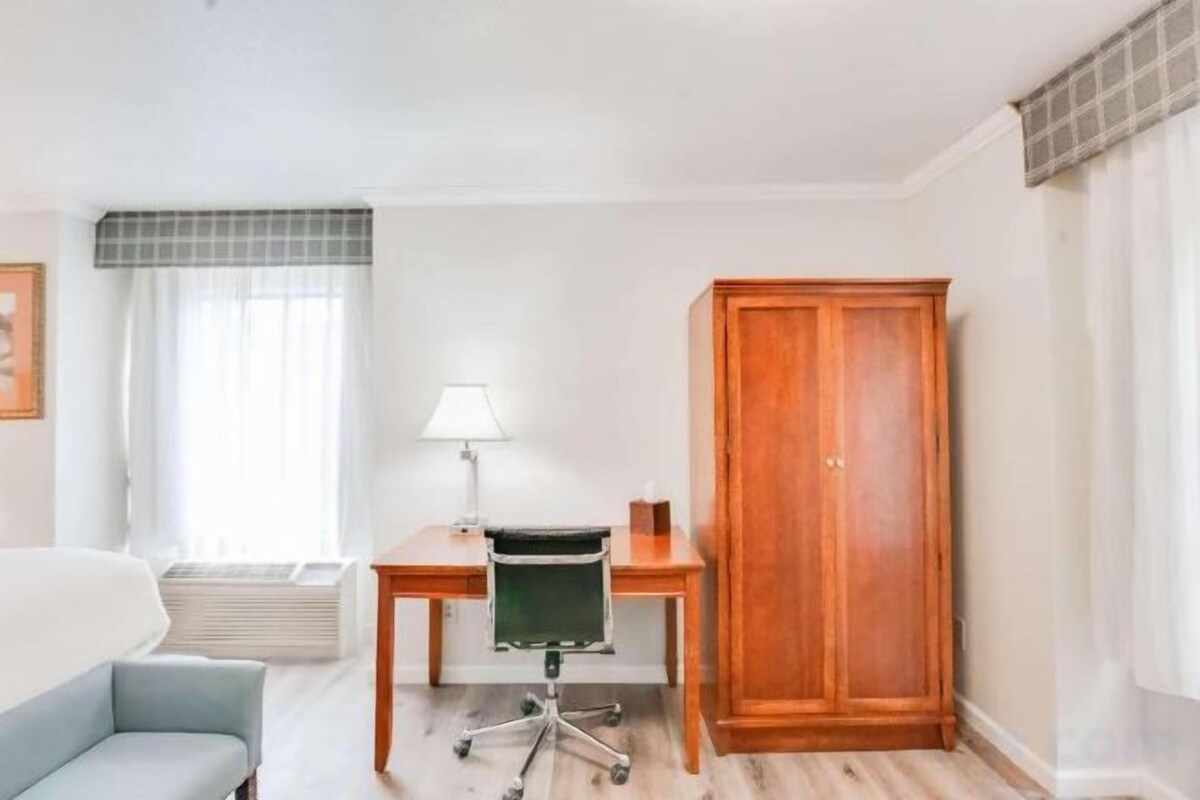 A hotel room with a bed, a desk with a chair, a wardrobe, and a window with curtains.
