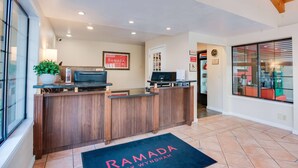 A reception desk with a wooden finish, a framed 'Ramada' sign, a potted plant, and a black mat with 'Ramada Wyndham' on it. - Ramada by Wyndham Mountain View (Mountain View)